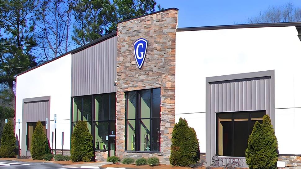 Das moderne Industriegebäude der Grace Academy mit Steinfassade und großem blauen Schild vor Bäumen und klarem Himmel.