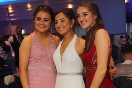 Drei Schülerinnen der Grange Post Primary School posieren gemeinsam in einer festlichen Umgebung mit anderen Personen im Hintergrund.