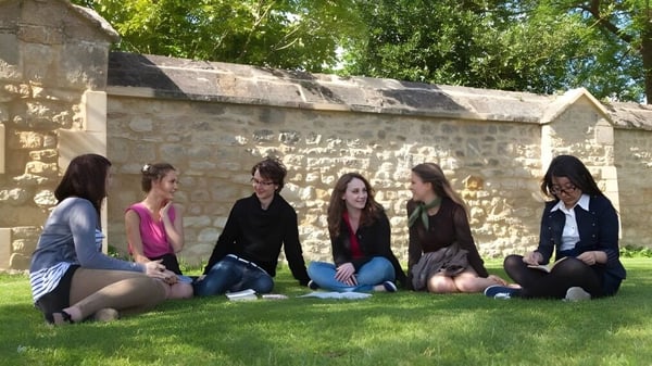 Eine Gruppe von Studentinnen sitzt auf der Wiese vor einer Steinmauer auf dem Campus von Greene's College Oxford.