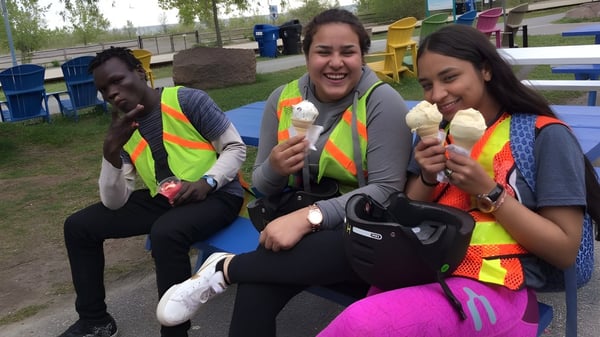 Drei Schülerinnen und Schüler der Greenwood Secondary School sitzen im Pausenhof und essen Eis vor einem bunten Spielplatz.