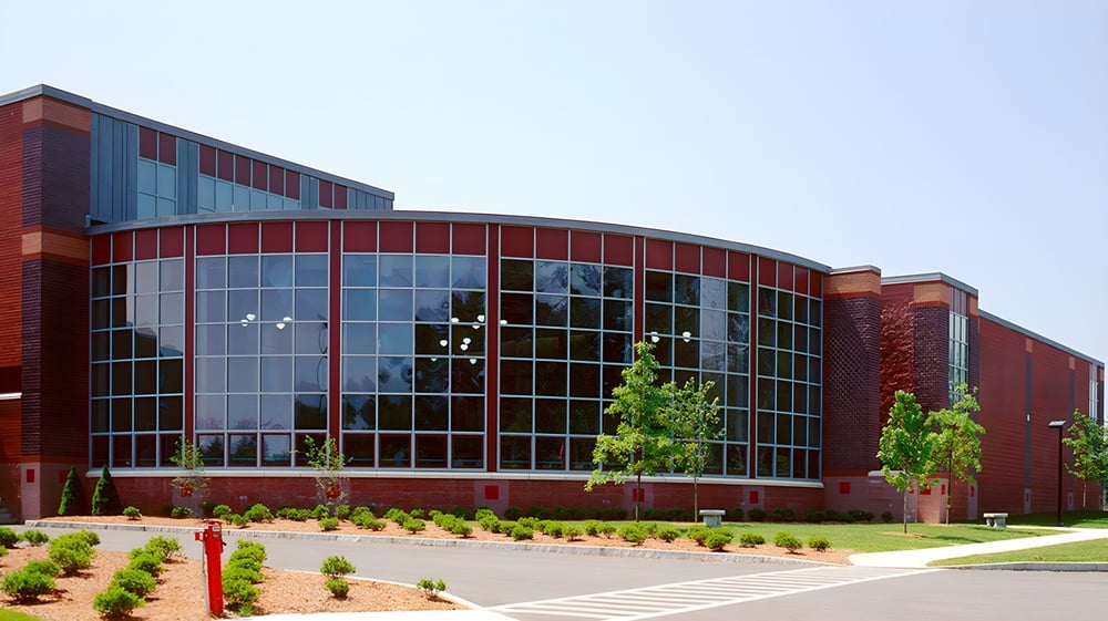 Das moderne mehrstöckige Gebäude der Groton-Dunstable Regional High School mit Glasfassade und rotem Backstein ist von Bäumen und Pflanzen umgeben.