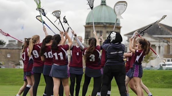 Eine Gruppe junger Athletinnen mit Lacrosse-Schlägern steht vor einem historischen Gebäude auf dem Campus der Haileybury School.
