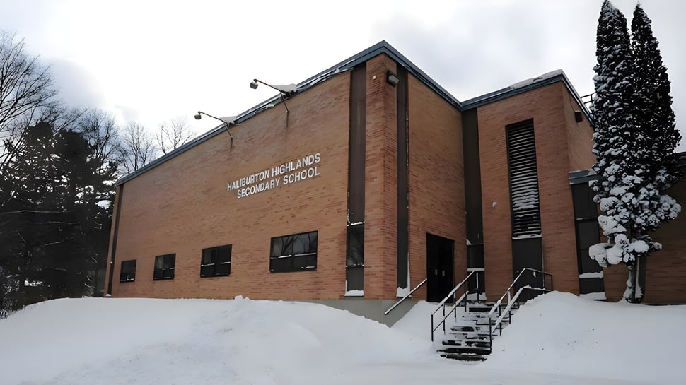 Das Schulgebäude der Haliburton Highlands School steht in einer verschneiten Landschaft mit sichtbaren Kiefern im Hintergrund.