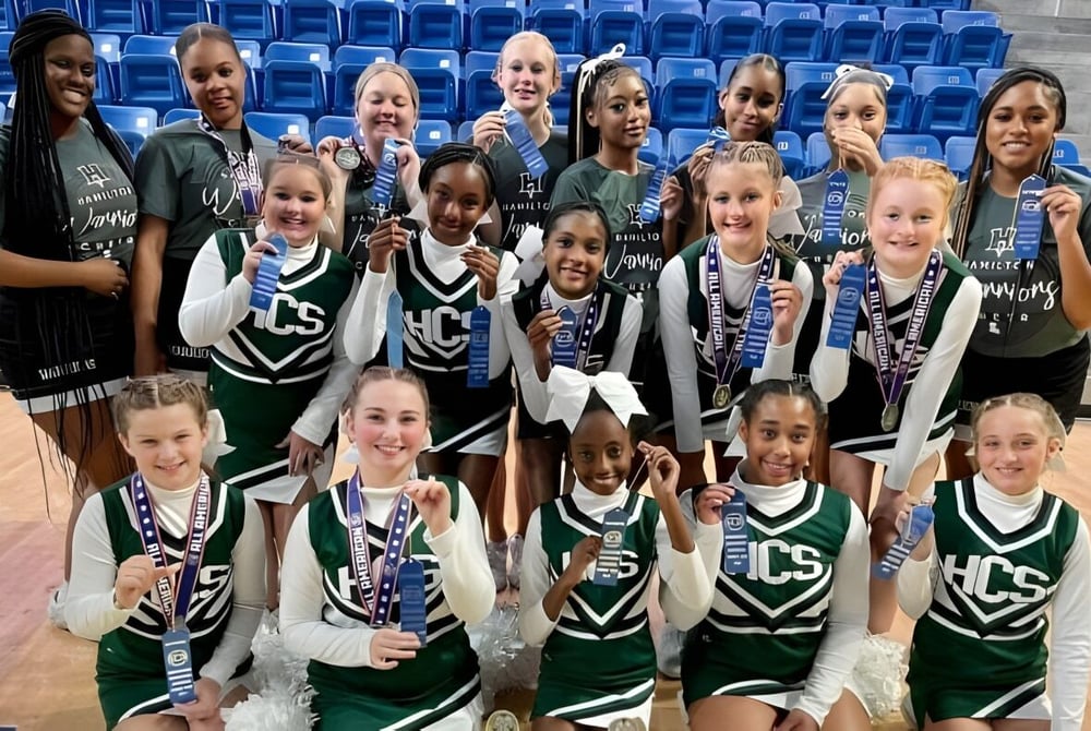 Eine Gruppe junger Basketballspielerinnen posiert auf dem Spielfeld der Hamilton Christian Academy mit blauen Tribünen im Hintergrund.