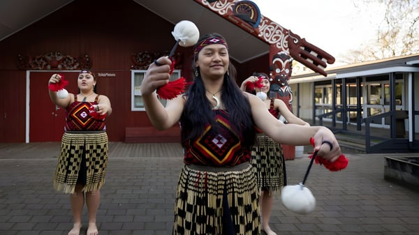 Zwei Schülerinnen der Hamilton Girl's High School tragen traditionelle Maori-Kleidung vor einem Gebäude mit markanter Architektur.
