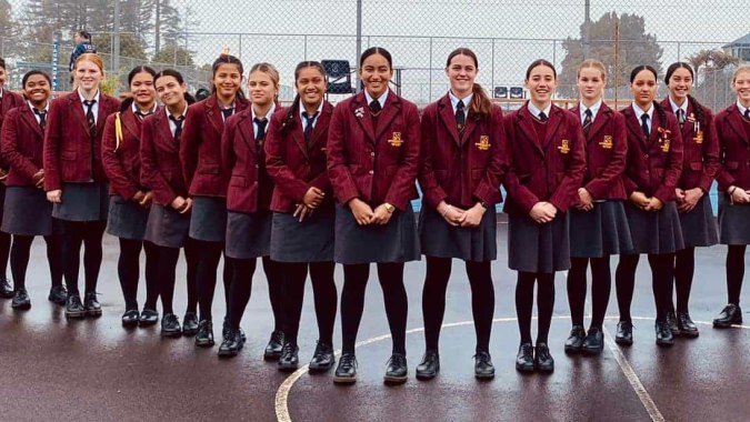 Eine Gruppe Schülerinnen in der Uniform der Hamilton Girl's High School steht zusammen im Freien vor Bäumen und Gebäuden.