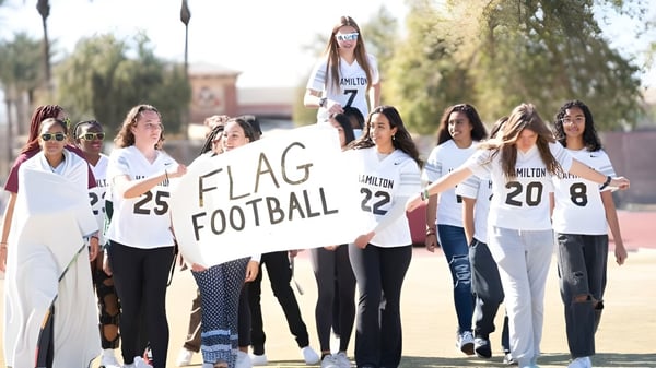 Eine Gruppe Schülerinnen der Hamilton High School steht mit einem Flag Football Schild vor dem Schulcampus.