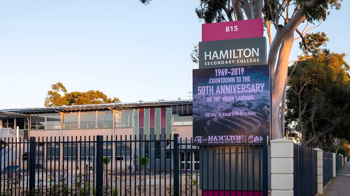 Der Eingang des Hamilton Secondary College mit einem großen Schild zur 50-Jahr-Feier und Palmen im Hintergrund.