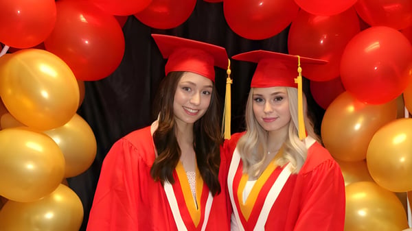 Zwei Absolventinnen der Hammarskjold High School posieren in ihren Abschlusskleidern vor roten und goldenen Luftballons.