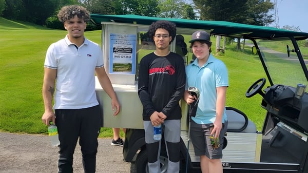 Drei Schüler der Harmony Christian School stehen vor einem Golfwagen auf einem Grasfeld mit Bäumen im Hintergrund.