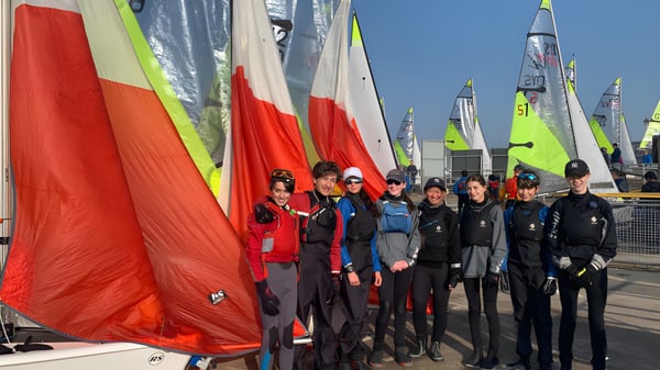 Eine Gruppe Schüler der Harrow School steht in bunter Segelausrüstung vor einer Reihe von Segelbooten mit roten und grünen Segeln.