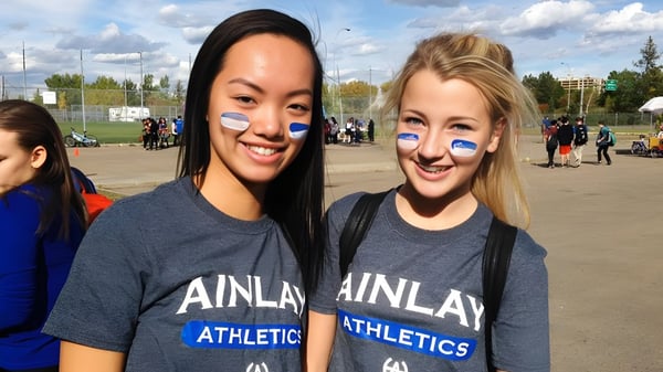 Zwei Schülerinnen der Harry Ainlay High School stehen lächelnd vor einem Sportfeld mit weiteren Personen im Hintergrund.
