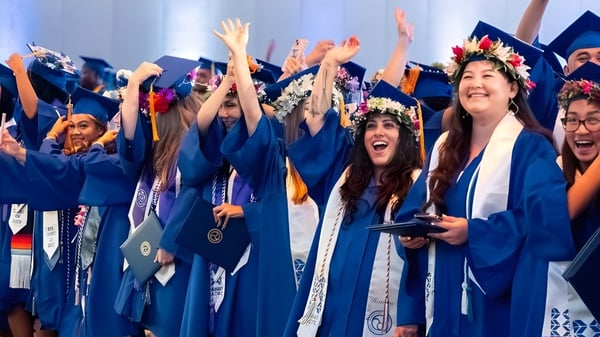 Eine Gruppe von Absolventinnen und Absolventen der Hawai'i Pacific University feiert mit erhobenen Armen in blauen Talaren und mit Blumenkränzen.