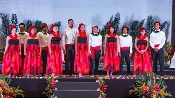 Schüler der Hawaiian Mission Academy stehen in roten und weißen Kostümen auf einer Bühne mit tropischer Kulisse.