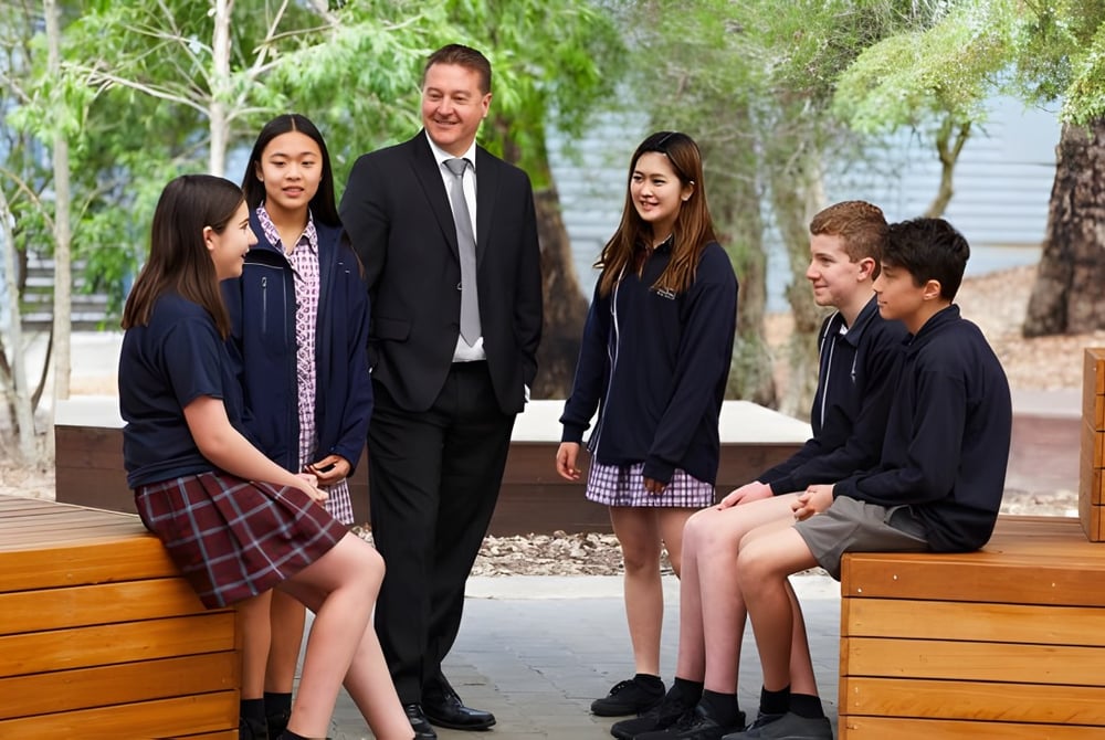 Eine Gruppe aus Schülern und einem Mann im Anzug steht auf der Holzterrasse der Heathfield High School umgeben von grüner Pflanzenwelt.