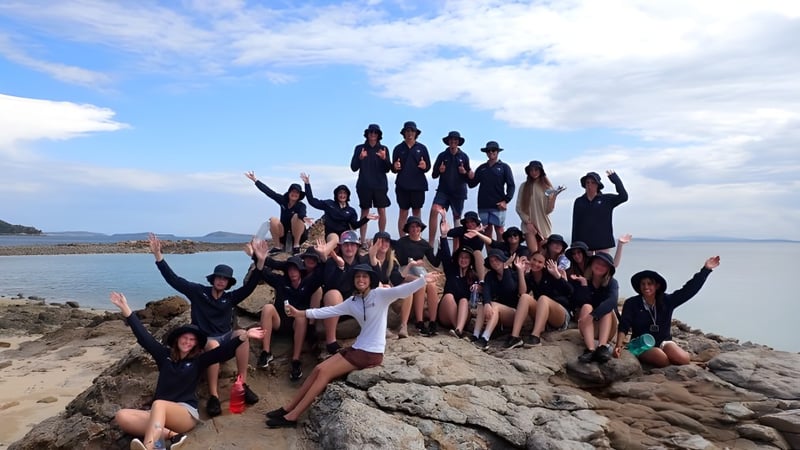 Eine Gruppe Schülerinnen und Schüler der Helensvale State High School posiert an einem felsigen Strand mit Bergen und blauem Himmel im Hintergrund.