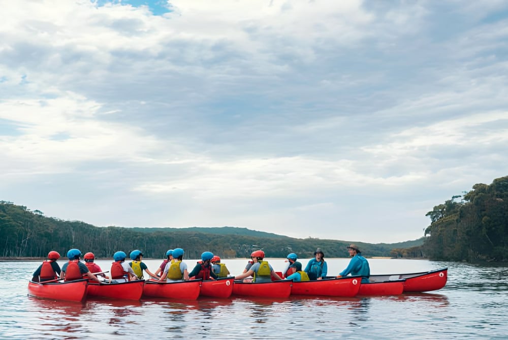 Schüler der Helensvale State High School paddeln in bunten Schwimmwesten mit einem roten Kanu auf einem See, umgeben von grünen Hügeln.