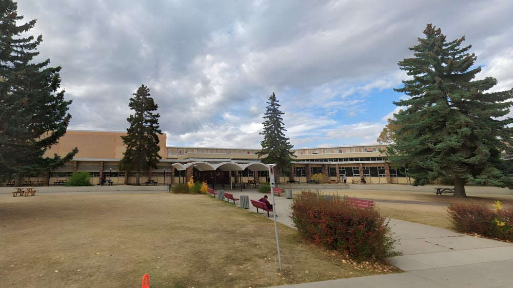 Das mehrstöckige Hauptgebäude der Henry Wise Wood High School mit überdachtem Eingang und begrüntem Vordergrund unter bewölktem Himmel.