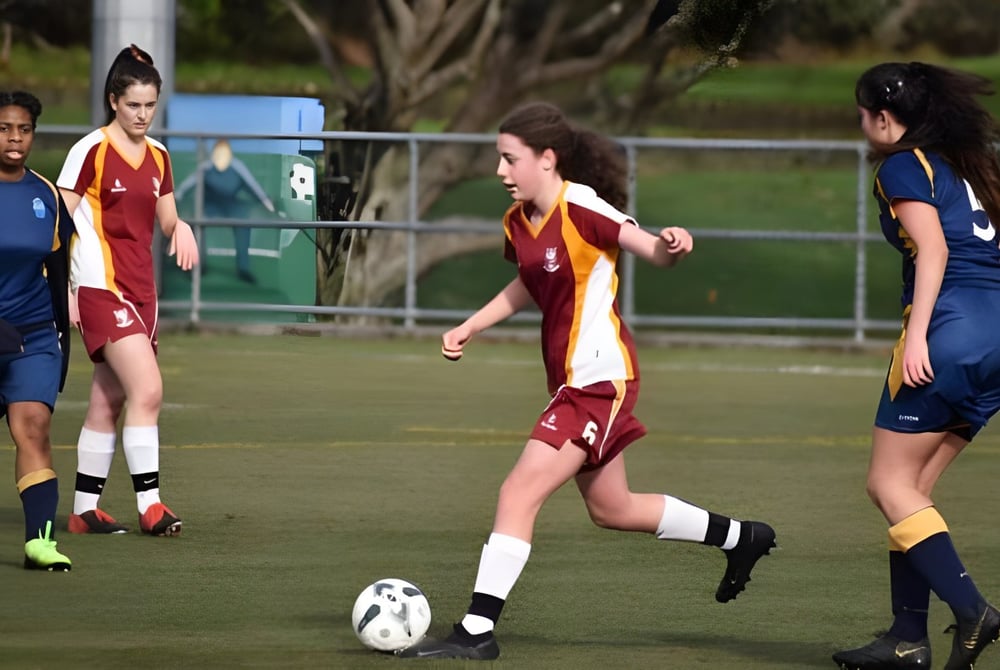 Schülerinnen vom Heretaunga College spielen ein Fußballspiel auf dem Rasenplatz mit Gebäuden im Hintergrund.