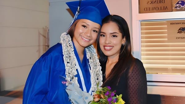 Zwei Schülerinnen der Heritage High School stehen gemeinsam in ihren Abschlussgewändern in einem Raum mit Fenster und Dekoration.