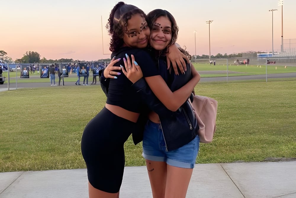 Zwei Schülerinnen umarmen sich auf einer Wiese vor dem Sportstadion der Heritage High School.