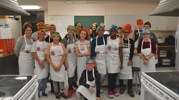 Schüler der Heritage Regional High School tragen weiße Schürzen und arbeiten in einer Schulküche mit Kochgeräten.