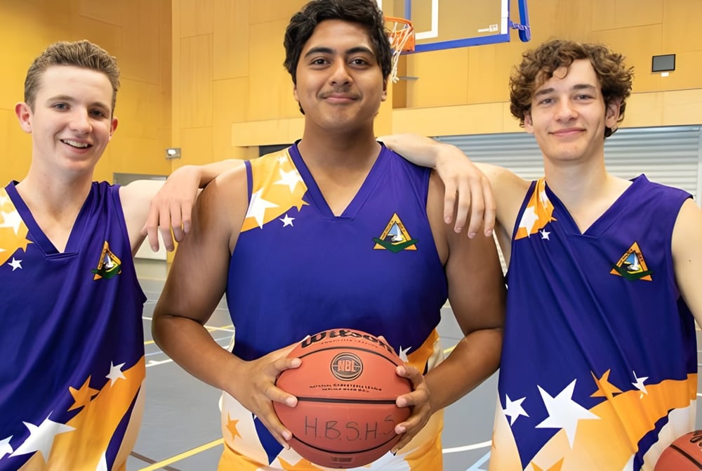 Drei Schüler der Hervey Bay State High School stehen in Basketballuniform in einer Sporthalle und halten einen Basketball.