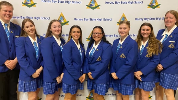 Eine Gruppe Schüler der Hervey Bay State High School steht vor einem Hintergrund mit dem Schulnamen und Logo.