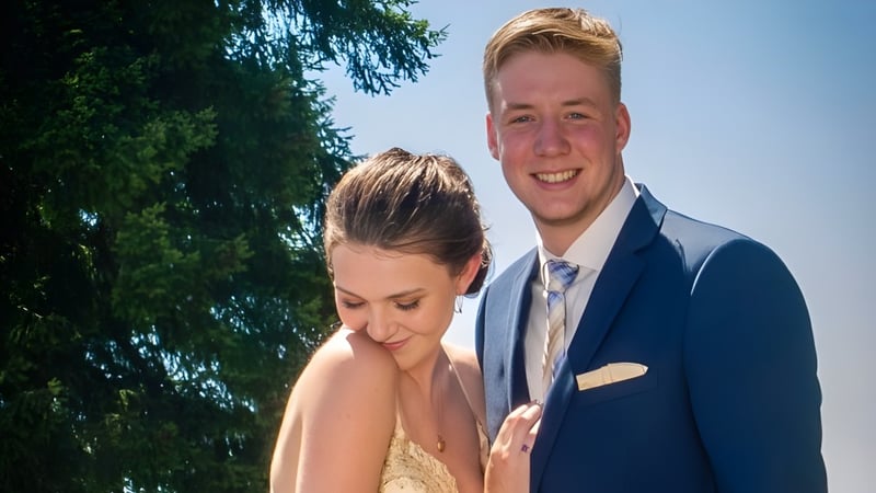 Ein Paar in formeller Kleidung steht im grünen Außenbereich der Highland Secondary School mit Bäumen im Hintergrund.
