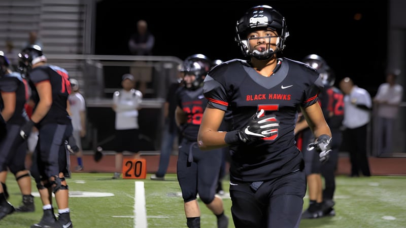Ein Footballspieler in schwarzer und roter Uniform steht auf dem Spielfeld des Higley Unified School Districts mit weiteren Spielern und Zuschauern im Hintergrund.