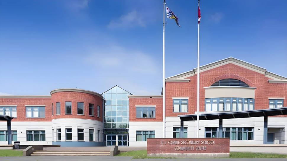 Das moderne mehrstöckige Backsteingebäude der H.J. Cambie Secondary School mit Glas-Eingang und Flaggen vor blauem Himmel.