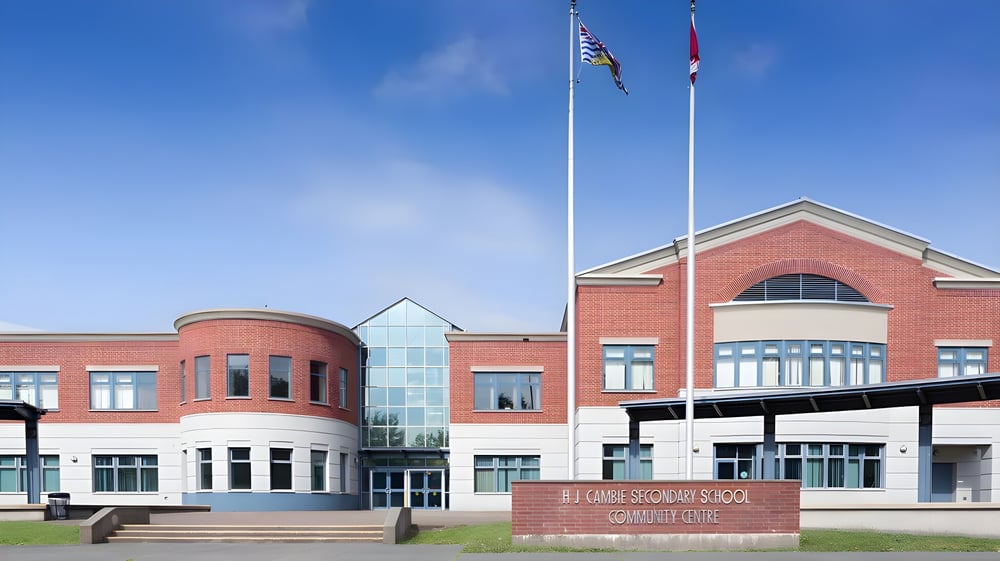 Das moderne mehrstöckige Backsteingebäude der H.J. Cambie Secondary School mit Glas-Eingang und Flaggen vor blauem Himmel.