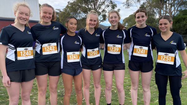 Eine Gruppe junger Athletinnen steht auf dem Campus der Hobart City High School vor einem bewaldeten Hintergrund.