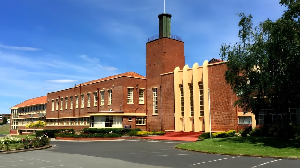 Das große Backsteingebäude mit hohem Schornstein auf dem Gelände der Hobart City High School ist von Bäumen und einer Einfahrt umgeben.