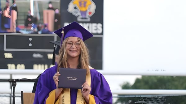 Eine Schülerin der Hobart City High School trägt eine violette Abschlussrobe und hält ihr Diplom vor einem Universitätslogo-Hintergrund.