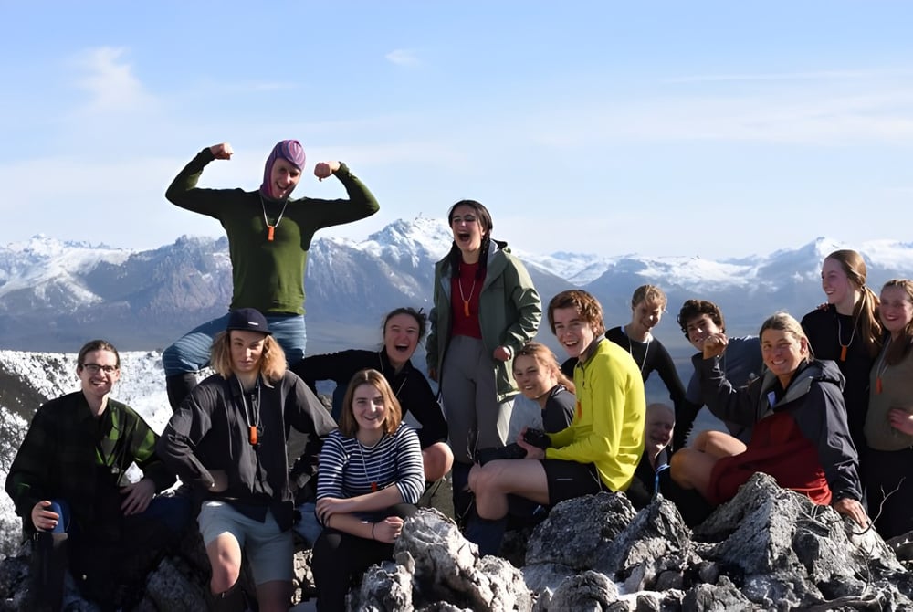Eine Gruppe von Schülerinnen und Schülern der Hobart City High School posiert auf einem Felsen mit schneebedeckten Bergen im Hintergrund.