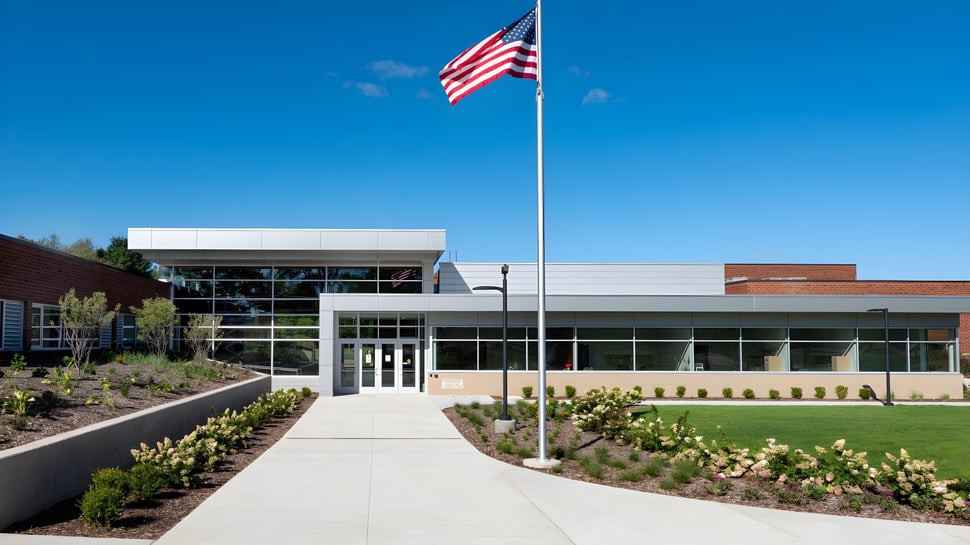 Das moderne Glasgebäude der Holland Christian Schools ist von einem gepflegten Garten mit verschiedenen Pflanzen und einem gepflasterten Weg umgeben.