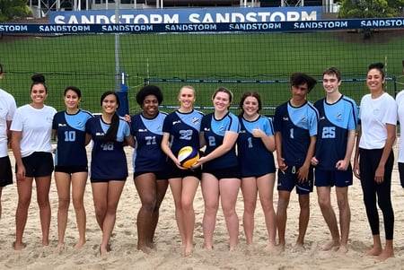 Schüler der Holland Park State High School stehen gemeinsam mit Volleyball auf einem Sandstrand vor dem Sandstorm-Schild.