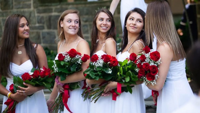 Eine Gruppe junger Frauen in weißen Kleidern hält Rosensträuße vor einer Steinmauer auf dem Gelände der Holy Child Secondary School.