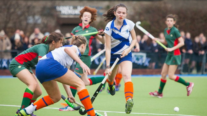 Schülerinnen der Holy Child Secondary School spielen ein Feldhockeyspiel auf einem Spielfeld mit Zuschauern im Hintergrund.