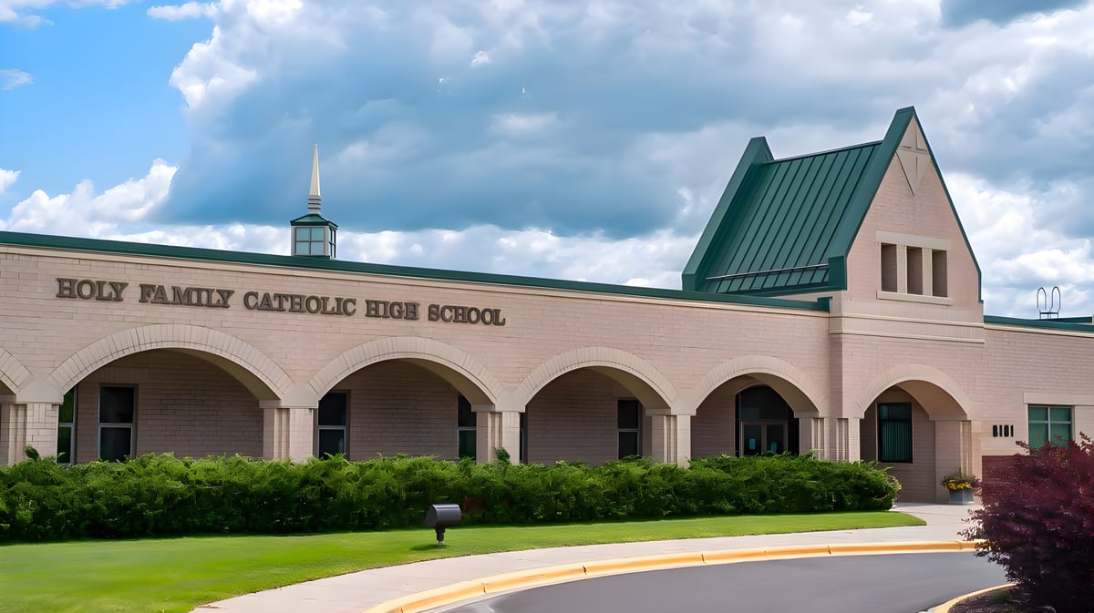 Das große Backsteingebäude der Holy Family Catholic High School mit Bogenportal und grünem Turmdach ist von Grün und bewölktem Himmel umgeben.