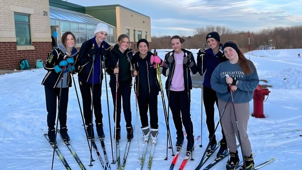 Eine Gruppe von Schülerinnen und Schülern der Holy Family Catholic High School steht in Winterkleidung auf Skiern im Schnee vor einem Gebäude.