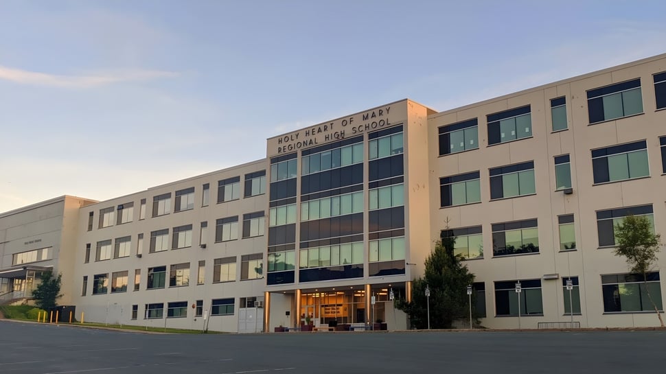 Das mehrstöckige Gebäude mit Glasfenstern der Holy Heart of Mary High School ist von Bäumen umgeben und hat einen gepflasterten Parkplatz im Vordergrund.