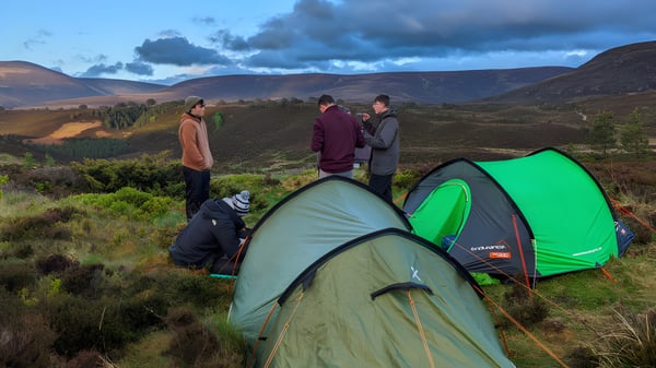 Eine Gruppe von Schülern der Holy Rood High School beim Camping in einer bergigen Landschaft mit Zelten im Vordergrund.