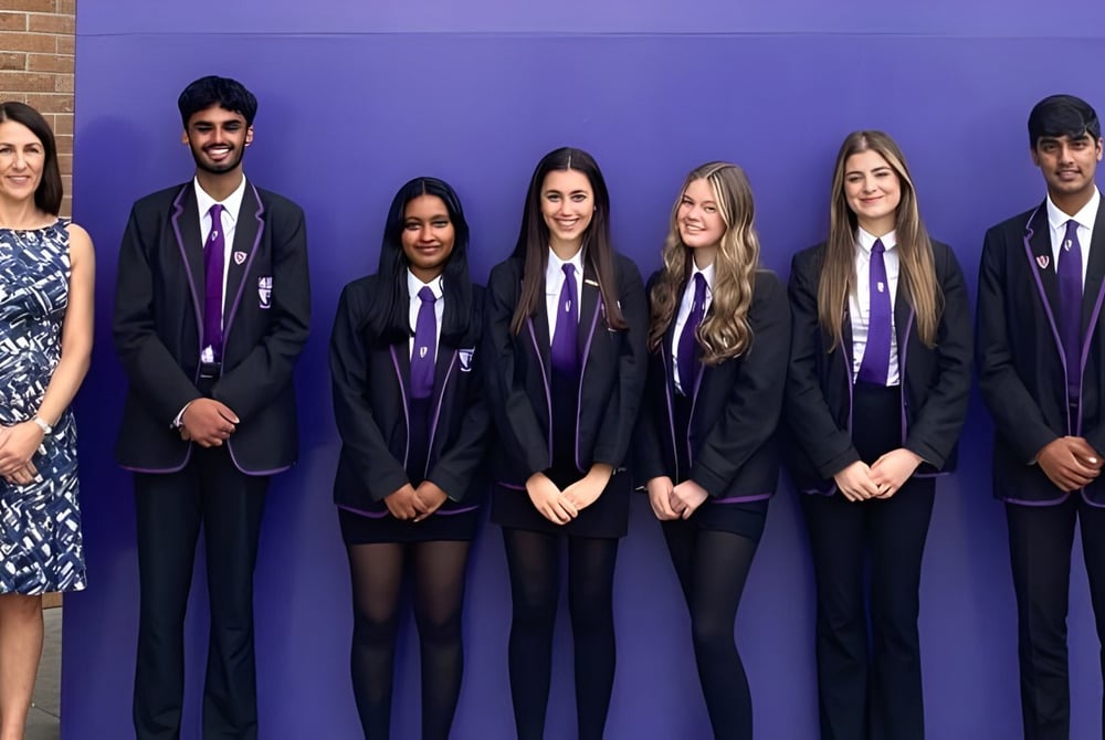 Eine Gruppe Schülerinnen und Schüler der Holy Rood High School steht in formeller Schuluniform vor einer lila Wand mit einer Frau im gemusterten Kleid.