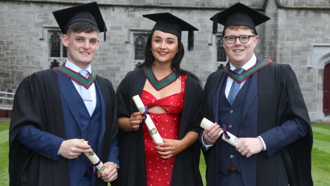 Drei Absolventen in Talaren mit ihren Diplomen vor einem Steingebäude auf dem Campus des Holy Rosary College.