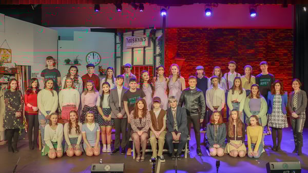 Eine Gruppe von Schülerinnen und Schülern steht auf der Bühne des Holy Rosary College vor einer bunten Beleuchtung und einer Ziegelsteinwand.