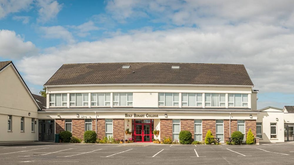Das zweigeschossige Backsteingebäude des Holy Rosary College mit rotem Ziegeldach steht vor einem bewölkten blauen Himmel.