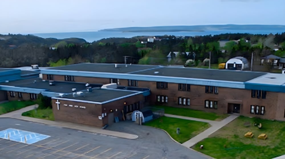 Das mehrstöckige Backsteingebäude der Holy Spirit High School steht auf einem grasbewachsenen Campus mit Blick auf ein Gewässer.