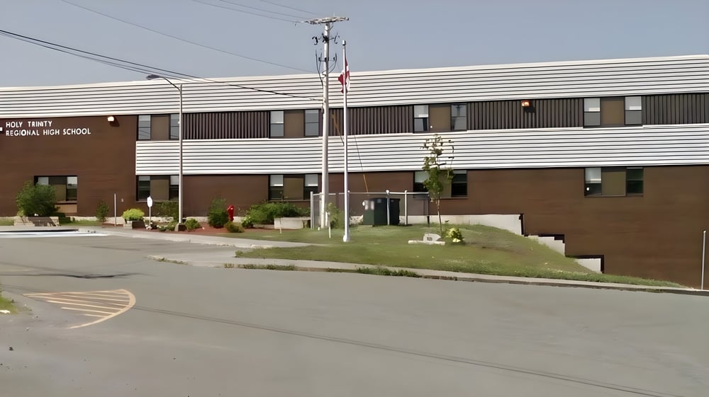 Das mehrstöckige Gebäude der Holy Trinity High School mit brauner Fassade steht umgeben von einer Grünfläche und einer Straße im Vordergrund.